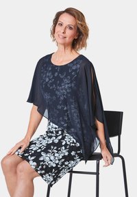 Flüssige marineblaue Bluse mit floralem Muster, kombiniert mit einem schwarzen Rock, der ein weißes Blumenmuster aufweist. Model sitzt auf einem schwarzen Stuhl.