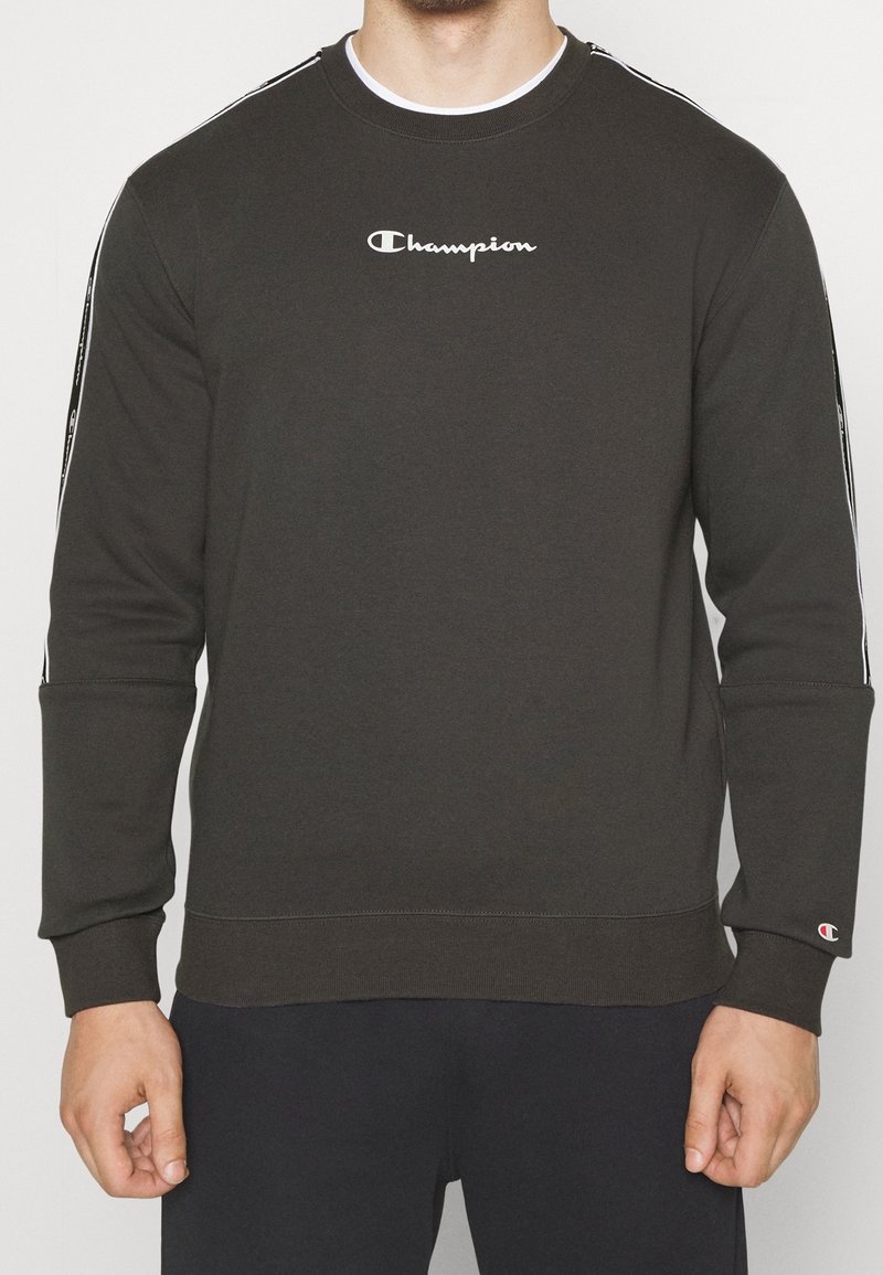 Uomo che indossa un felpa nera Champion con logo bianco sul petto e strisce di marca sulle maniche, in piedi contro uno sfondo semplice.