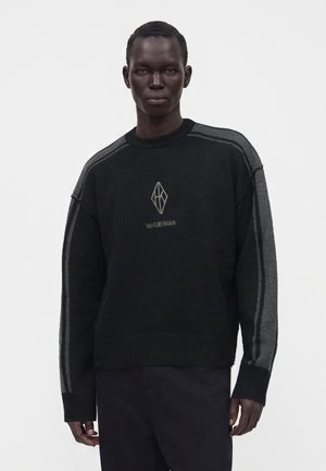 Homme portant un pull noir et gris à manches longues avec un logo géométrique et du texte sur la poitrine, debout devant un fond blanc uni.