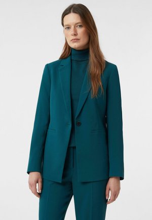 Woman with long brown hair wearing a teal turtleneck sweater, teal blazer, and matching teal trousers against a plain background.