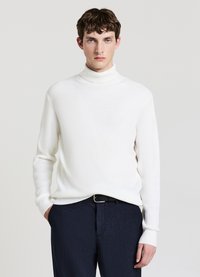 Maglione bianco a coste con collo alto e maniche lunghe, abbinato a pantaloni blu navy con trama e cintura nera. Design e vestibilità semplici e moderni.