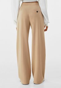 Pantaloni a gamba larga in morbido tessuto beige, caratterizzati da una superficie liscia, vita alta e una tasca posteriore con un dettaglio in bottone nero.