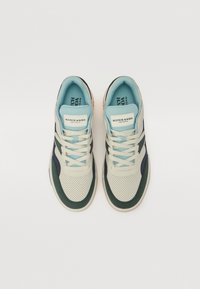 Stylish sneakers featuring a cream perforated upper, green and dark blue accents, and light blue laces. Rubber sole with logo branding.