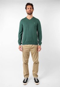 Pull vert en V en matière tricotée, associé à un pantalon chino beige et des baskets noires. Le pull a des manches longues et un détail logo discret.