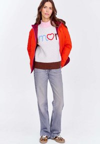 Kobieta ubrana w pomarańczową kurtkę na białym swetrze z napisem "amor", jasnoniebieskie dżinsy i sneakersy w lamparcim wzorze, stojąca na białym tle.