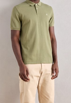 Man wearing olive green short-sleeve polo shirt with two buttons and beige pants, standing against a plain light gray background.