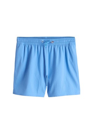 Młody mężczyzna w kremowym teksturowanym polo z krótkim rękawem, jasnoniebieskich szortach, czarnych sandałach na wsuwkę i okularach przeciwsłonecznych stojący przy teksturowanej beżowej ścianie.