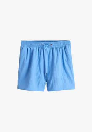 Jeune homme portant un polo crème texturé à manches courtes, un short bleu clair, des sandales noires et des lunettes de soleil, debout contre un mur beige texturé.