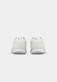 Paire de baskets basses blanches vues de dos, avec une semelle côtelée et de petites marques noires et beiges sur la languette du talon.