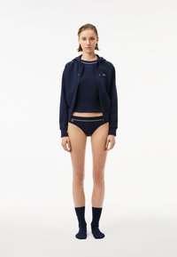 Mörkblå zip-up hoodie med dragsko, kombinerad med en croppad topp och matchande bikini, med kontrasterande vita detaljer och logotyp.