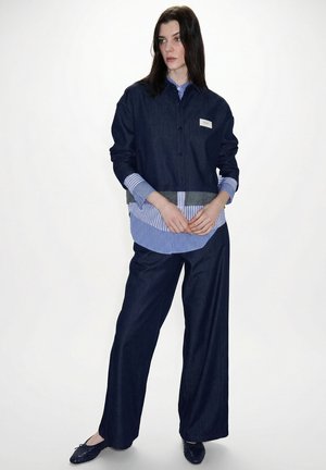 Donna che indossa una camicia a strati a righe blu sotto una giacca di denim scuro e pantaloni a gamba larga, in piedi davanti a uno sfondo bianco.