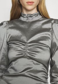 Top metallico grigio con un colletto alto arricciato, dettagli raccolti nella parte frontale e maniche a palloncino. Texture liscia con una leggera lucentezza.
