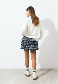 Pull blanc associé à une jupe plissée mini à carreaux bleu marine. Chaussures crème avec une semelle épaisse et des chaussettes rayées. Textures lisses et design superposé.