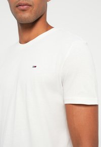 Uomo che indossa una semplice maglietta bianca a maniche corte con un piccolo logo rettangolare rosso, bianco e blu sul petto sinistro.
