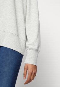 Sweat-shirt gris à coupe décontractée, avec des poignets côtelés et une texture douce, associé à un jean en denim foncé. Détail du poignet et de la main visible.