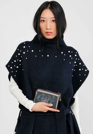 Poncho de lana azul marino con cuello alto y adornos de perlas, usado sobre una blusa de manga larga clara, sosteniendo una cartera azul con estampado.