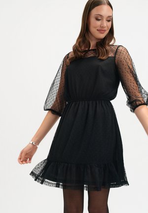 Czarna sukienka z siateczkową nakładką w grochy, krótkimi bufkowatymi rękawami i falbaną na dole. Posiada elastyczną talię, podkreślającą sylwetkę.