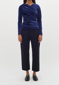 Top lungo con maniche a raglan in velluto blu navy e design avvolgente, abbinato a pantaloni neri a vita alta e larghi, con mocassini neri, indossato da un modello.