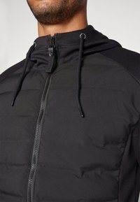 Svart zip-up jakke med kviltet tekstur, snøring på hette, og et elegant design med glatt stoff og kontrasterende skulderpanel.