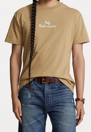 Tan cotton t-shirt with short sleeves, featuring white embroidered "Polo Ralph Lauren" text across the chest. Worn with blue jeans.