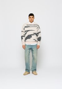 Puhas, krém színű pulóver fekete absztrakt mintázattal; laza szabással tervezték. Világoskék farmerral és bézs cipővel párosítva.