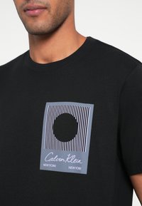 Melna kokvilnas T-krekls ar grafisku apdruku, tajā ir vertikālas svītras un aplis ar uzrakstiem "Calvin Klein" un "NEW YORK".