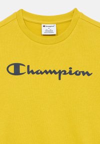 Κίτρινο βαμβακερό μπλουζάκι με μπλε σκούρο λογότυπο "Champion" στο στήθος και κλασικό σχέδιο με στρογγυλή λαιμόκοψη. Ομαλή υφή χωρίς σχέδια.