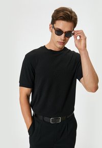 Sort kortærmet t-shirt med en tekstureret mønster, parret med mørke solbriller. Sort bælte båret, hænderne placeret nær solbrillerne.