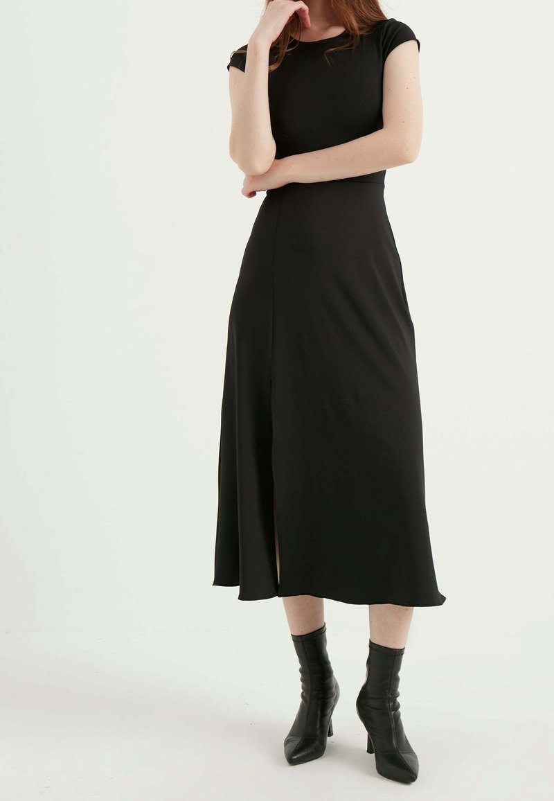 Sort midi-kjole med korte ærmer, rund halsudskæring og en slids foran. Kombineret med sorte ankelstøvler med hæl. Glat tekstur.