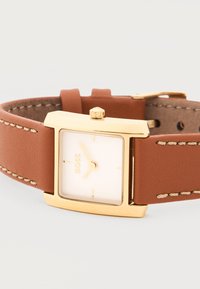 Ορθογώνιο χρυσό ρολόι με λευκό καντράν, μίνιμαλ σημάδια, λογότυπο BOSS και καφέ δερμάτινο λουρί με μπεζ ραφές.