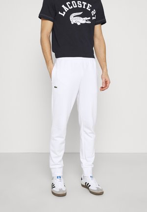 Man draagt witte Lacoste trainingsbroek, zwart Lacoste T-shirt met krokodillenlogo en witte Adidas sneakers met zwarte strepen.