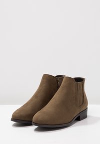 Botas de tobillo de gamuza marrón con cremallera lateral y paneles elásticos, que presentan un bajo tacón apilado y un diseño de punta redonda. Textura suave.