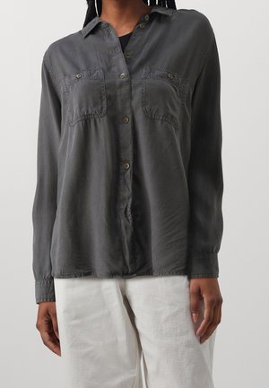 Vrouw draagt een donkergrijs, met knopen gesloten, longsleeve shirt met borstzakken en een witte broek, staand tegen een effen lichte achtergrond.