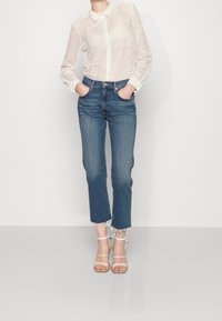 Blusa de encaje con cierre de botones y cuello; jeans azules con dobladillo deshilachado; sandalias blancas de tiras; la modelo posa con las manos en los bolsillos.