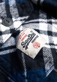 Sininen ja musta flanellikangas, jossa on valkoinen ruutukuvio; sisältää teksturoidun etiketin, jossa lukee "SuperDry" mustilla ja punaisilla yksityiskohdilla.
