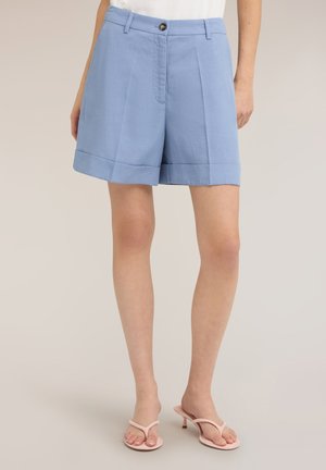Femme portant un short bleu clair cintré avec des plis à l'avant, des ourlets retournés et une fermeture à boutons, associé à des sandales à talons rose pâle.