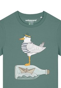 Groene shirt met een cartoonzeemeeuw in een gestreept shirt en hoed, die op een fles staat met een papieren boot en golven.