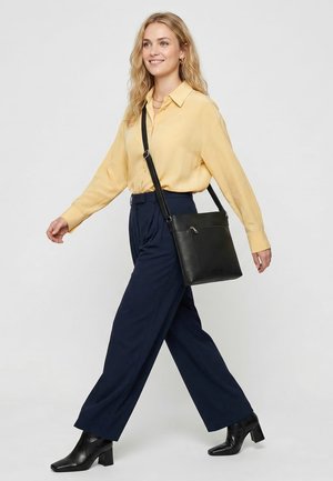 Woman walking wearing a yellow blouse, navy wide-leg pants, black heeled boots, and a black crossbody bag.