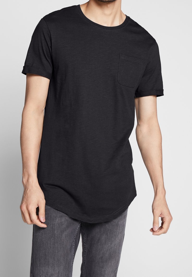 T-shirt en coton noir à manches courtes, avec un col rond et une poche poitrine. L'ourlet présente une légère courbure.
