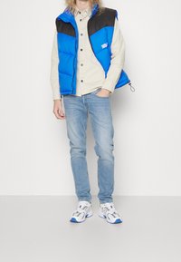 Person klädd i en blå och svart pufferväst, beige skjorta med knappar, ljusblå jeans och vit-blå sneakers som står mot en enkel vägg.