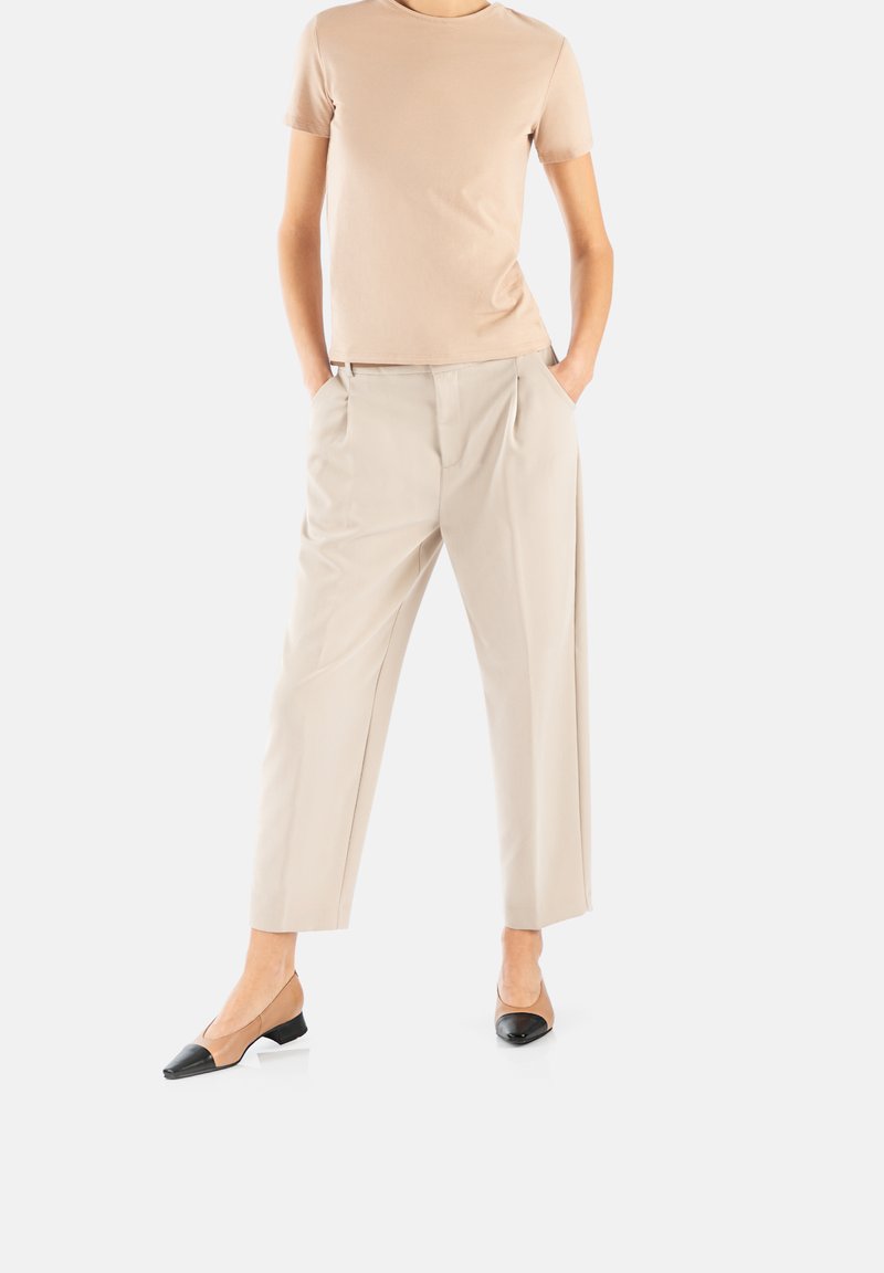 Beige Baumwoll-T-Shirt kombiniert mit gepolsterten, maßgeschneiderten cremefarbenen Hosen und schwarz-beigen, flachen Schuhen mit niedrigem Absatz, die eine strukturierte Silhouette schaffen.