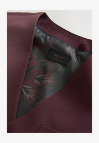 Blazer marrón mostrando el forro interior con estampado floral y etiqueta de la marca NEXT en negro con texto "Hecho en Bangladesh".