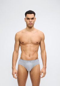 Άνδρας με αθλητική διάπλαση που φοράει ανοιχτό γκρι εσώρουχα Calvin Klein, στέκεται μπροστά σε απλό φωτεινό φόντο.