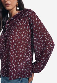 Blusa bordeaux con maniche lunghe e a sbuffo, caratterizzata da un collo arr gathered e piccoli motivi floreali blu su tutta la superficie. Tessuto leggero e texturizzato.
