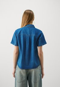 Blå denimskjorte med kort erm, krage-design og detalj med plissé bak. Stoffet er litt teksturert med en avslappet passform.