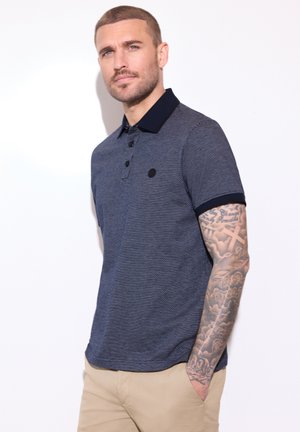 Homme aux cheveux courts et aux bras tatoués portant un polo texturé bleu marine et un pantalon beige, debout devant un fond blanc.