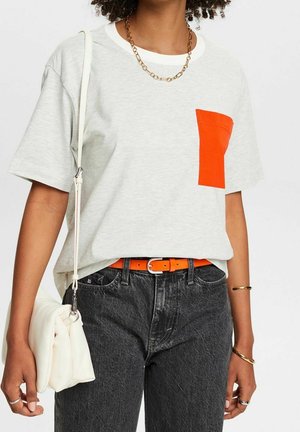 Personne portant un T-shirt gris clair avec une poche orange, un jean noir, une ceinture orange, un collier chaîne doré, des bracelets, des bagues, et tenant un sac à main blanc.