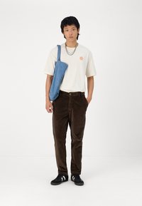 Lys beige t-skjorte med en oransje solgrafikk, brune kordfløyelsbukser og svarte joggesko med hvite striper, komplementert av en blå skulderveske.