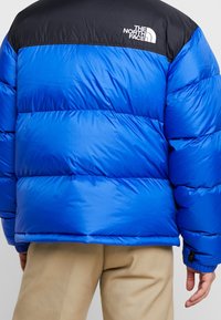 Doudoune bleue avec une section supérieure noire, présentant des coutures horizontales, un logo sur l'épaule, et une texture lisse et brillante.