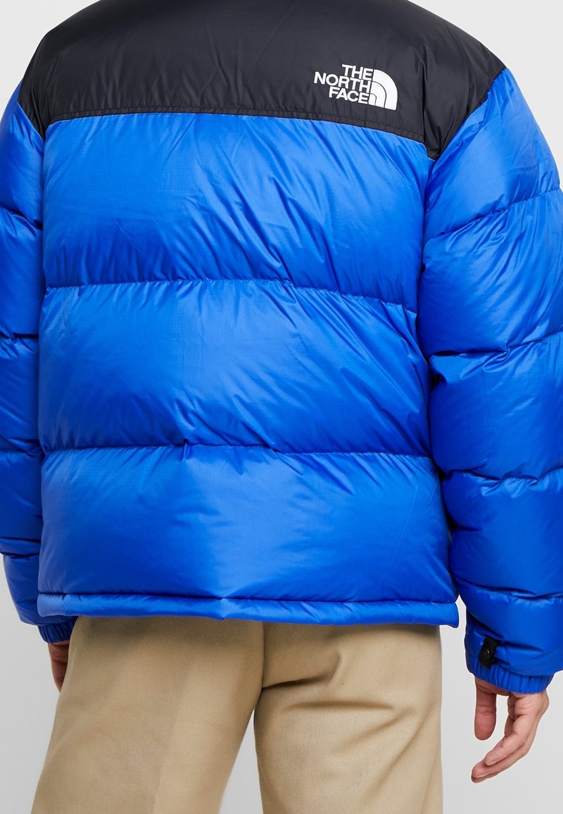 Doudoune bleue avec une section supérieure noire, présentant des coutures horizontales, un logo sur l'épaule, et une texture lisse et brillante.